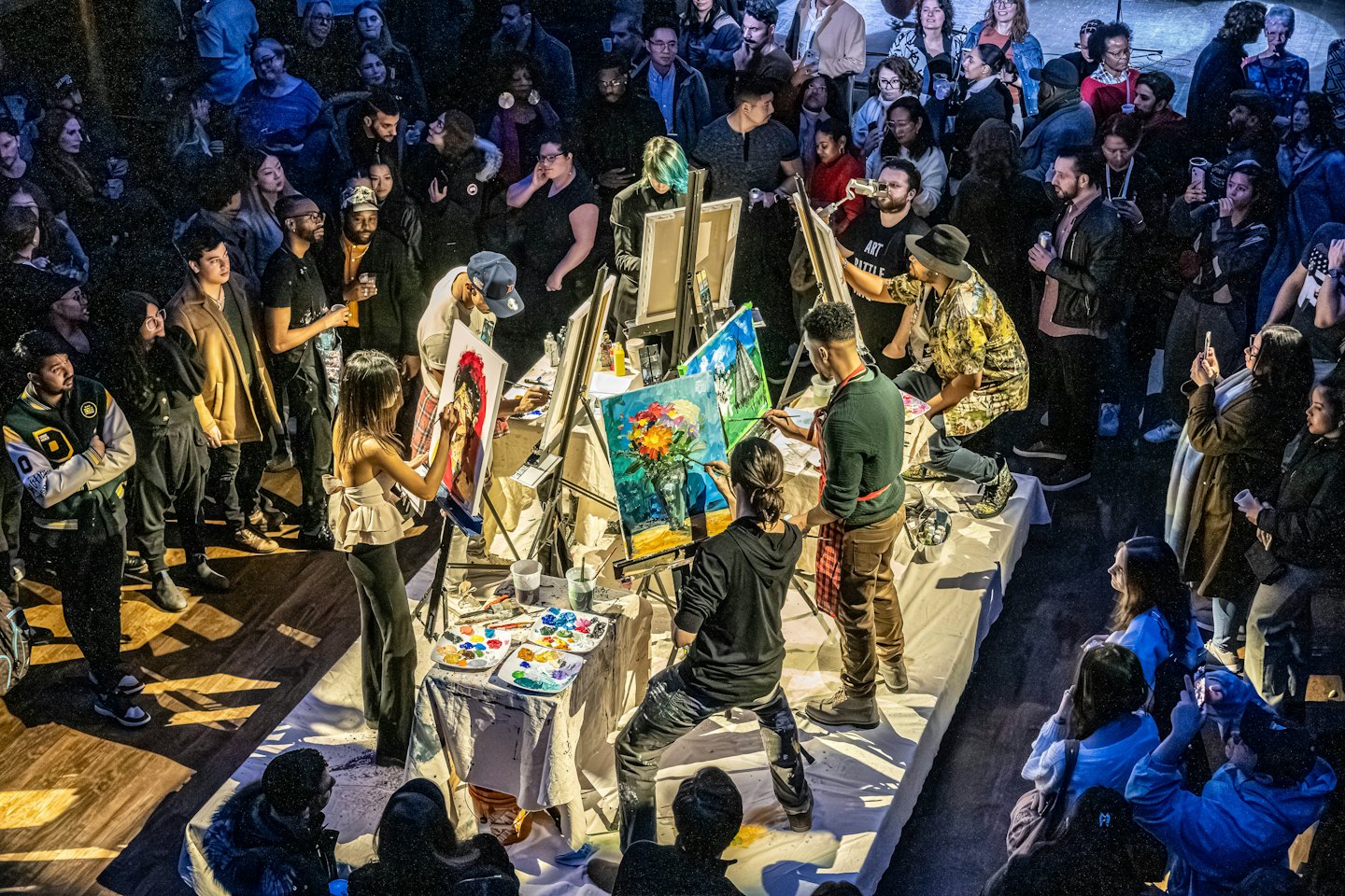 Artists painting at easels during an indoor competition with an audience.